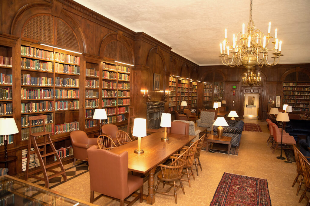  A large room with wood-paneled walls, a coffered ceiling, and a large fireplace. There are several tables and chairs in the room, as well as a large rug and a chandelier. The room is filled with bookshelves, and there is a librarian standing at a desk near the entrance.