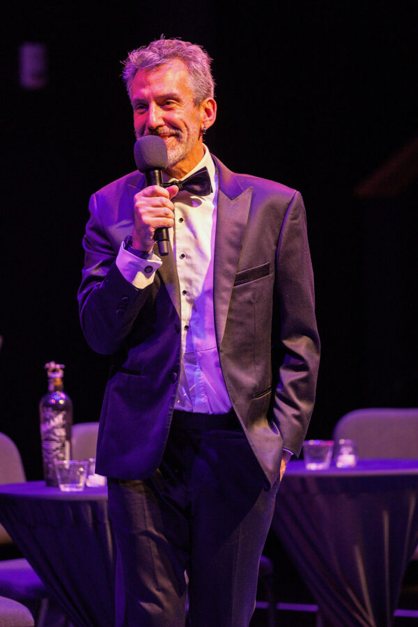 Bart Kuebler, dressed in a tuxedo, speaks into a microphone on a dark stage.