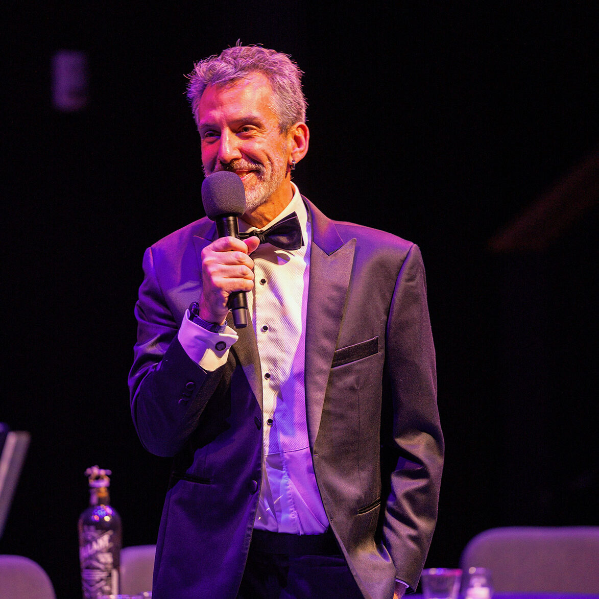 Bart Kuebler, dressed in a tuxedo, speaks into a microphone on a dark stage.