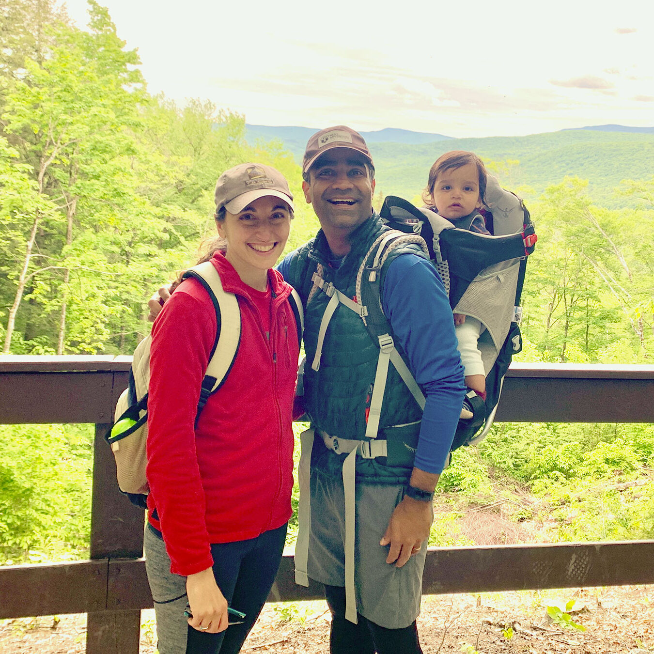 Gairik Sachdeva carries his child in a backpack and stands next to his wife on a bridge overlooking a forest and mountain vista.