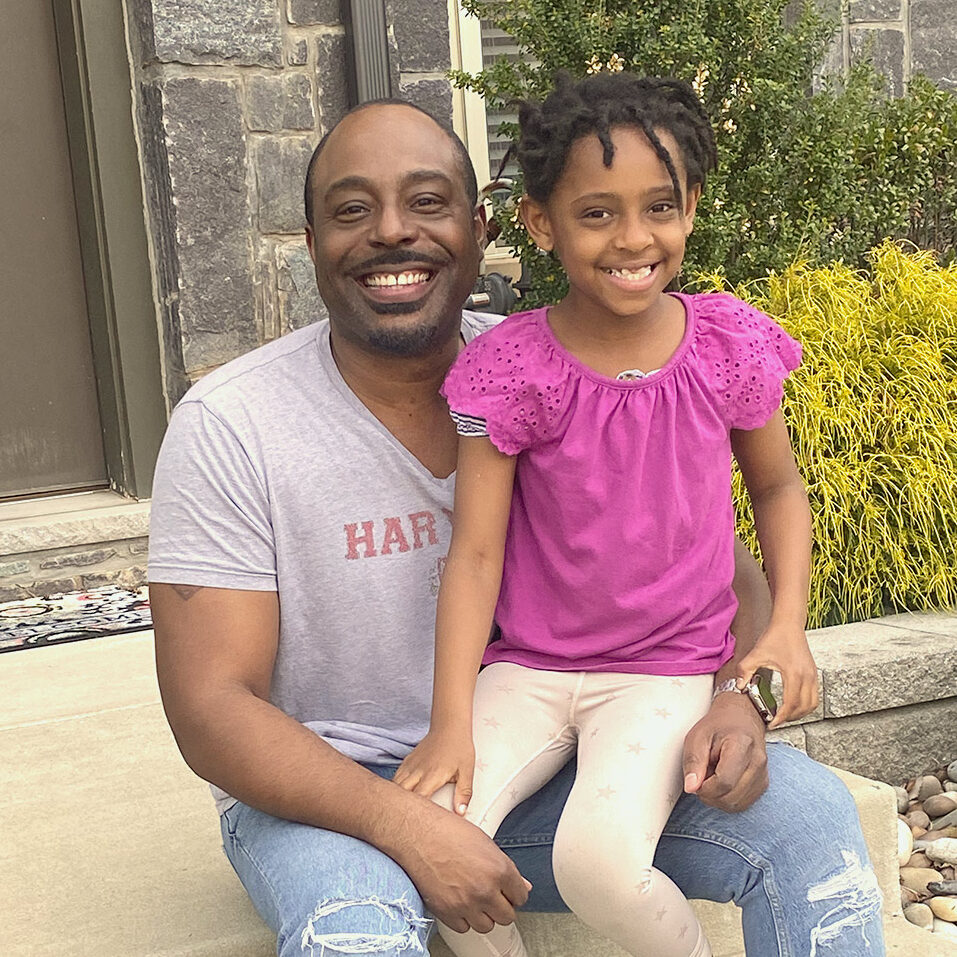 Kevin Simmons, in a Harvard t-shirt, sits on a doorstep with his daughter on his knee.