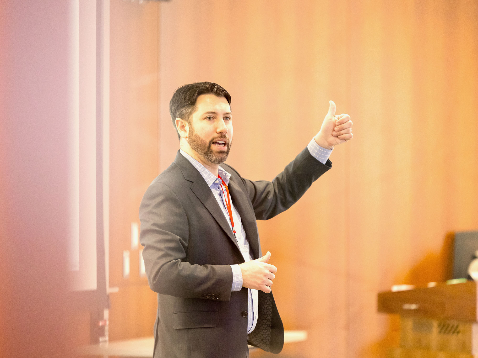 A man in a suit stands in front of a projection screen, gesturing with his hand as he presents to a classroom.