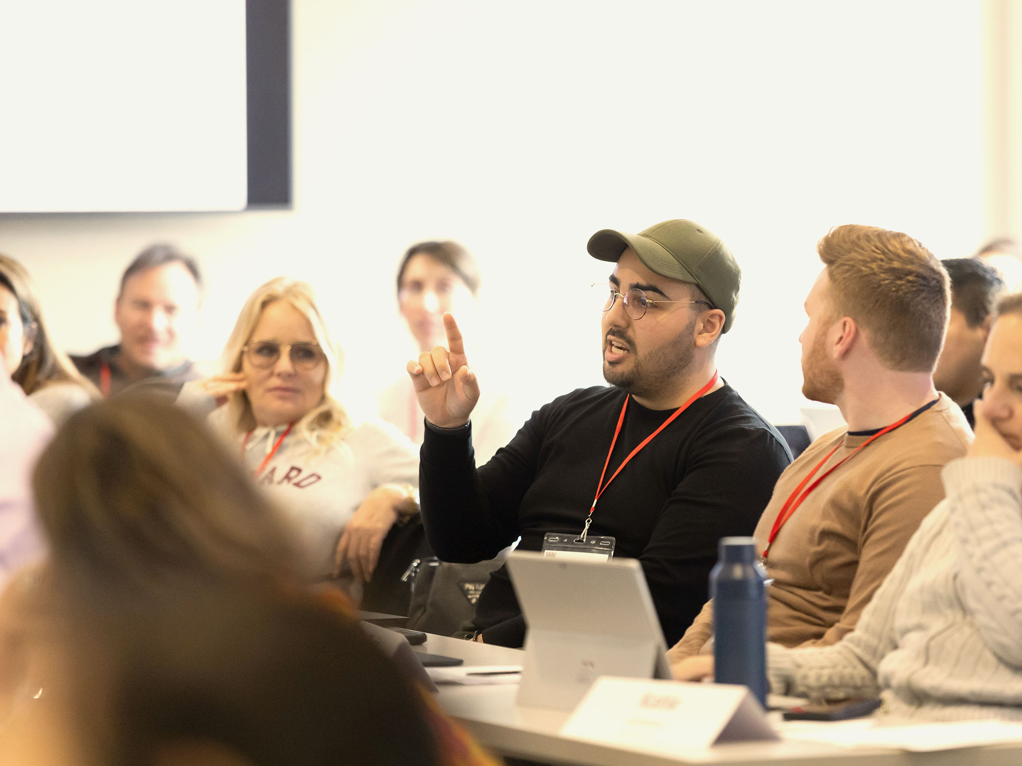 A man wearing a green cap gestures with one finger raised during a discussion, surrounded by peers.