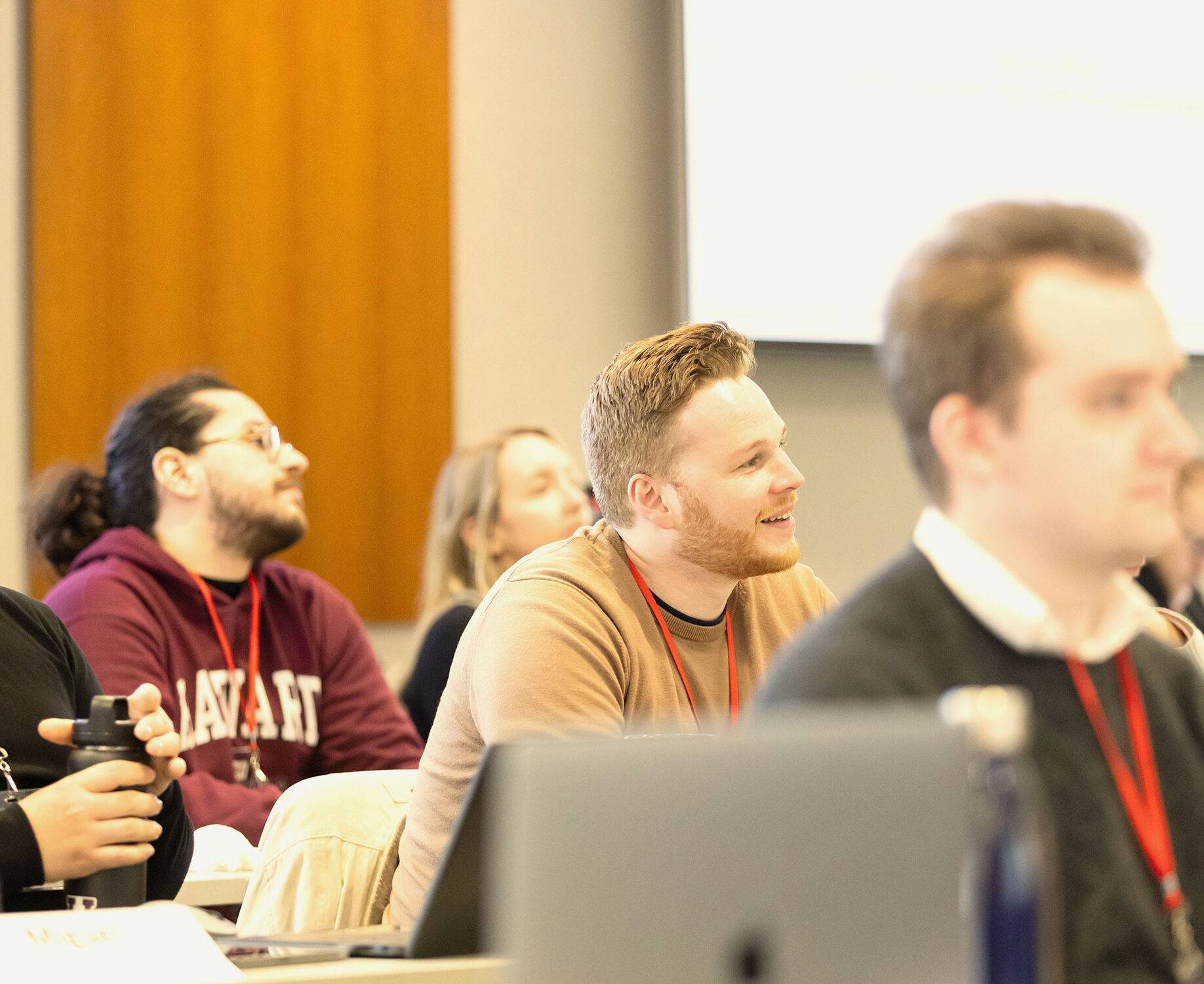 Classroom of students listening attentively, one man in a tan sweater smiles while facing forward.