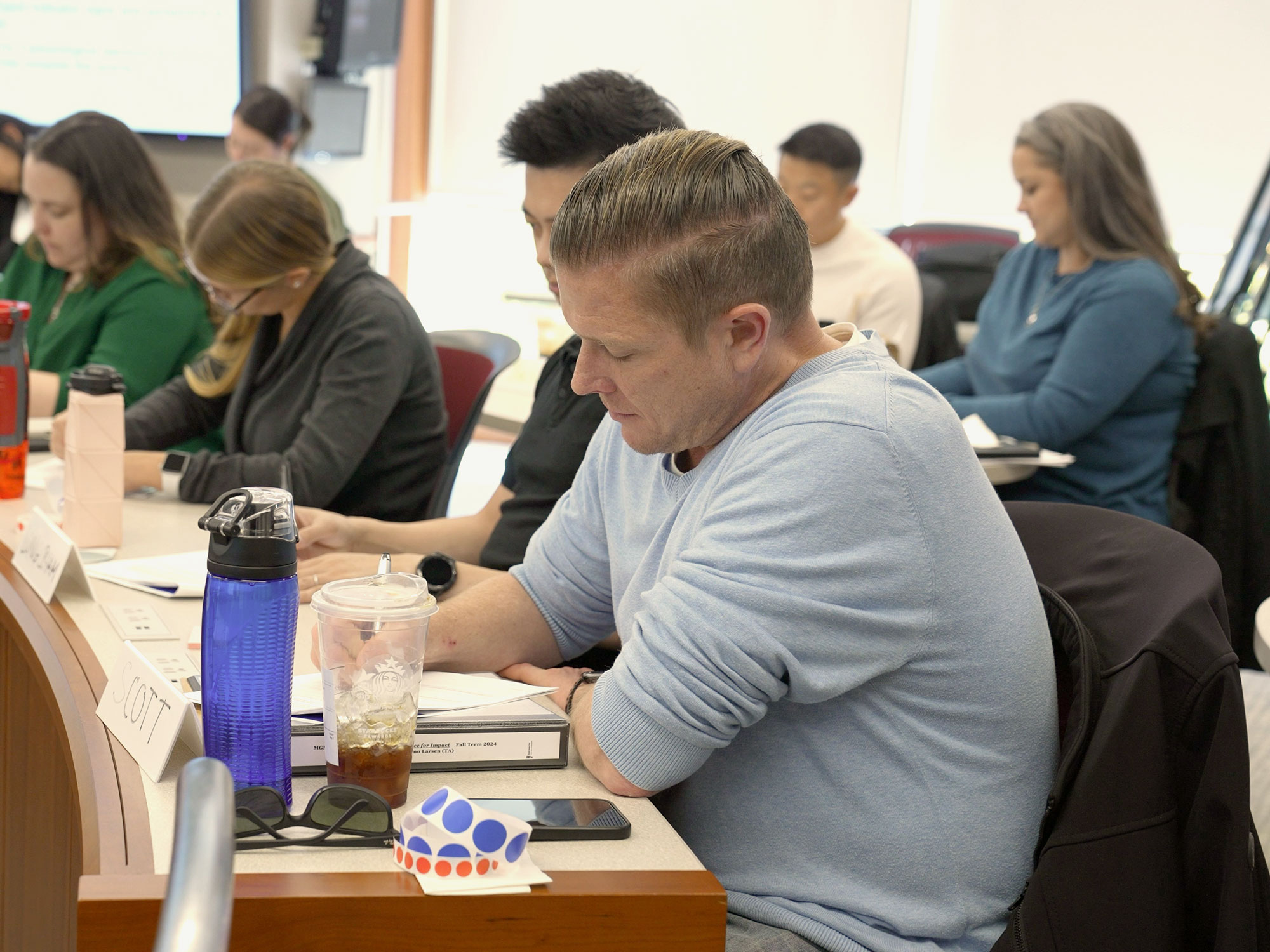 A man in a light blue sweater reads a document during class, with notebooks and drinks on the desk.