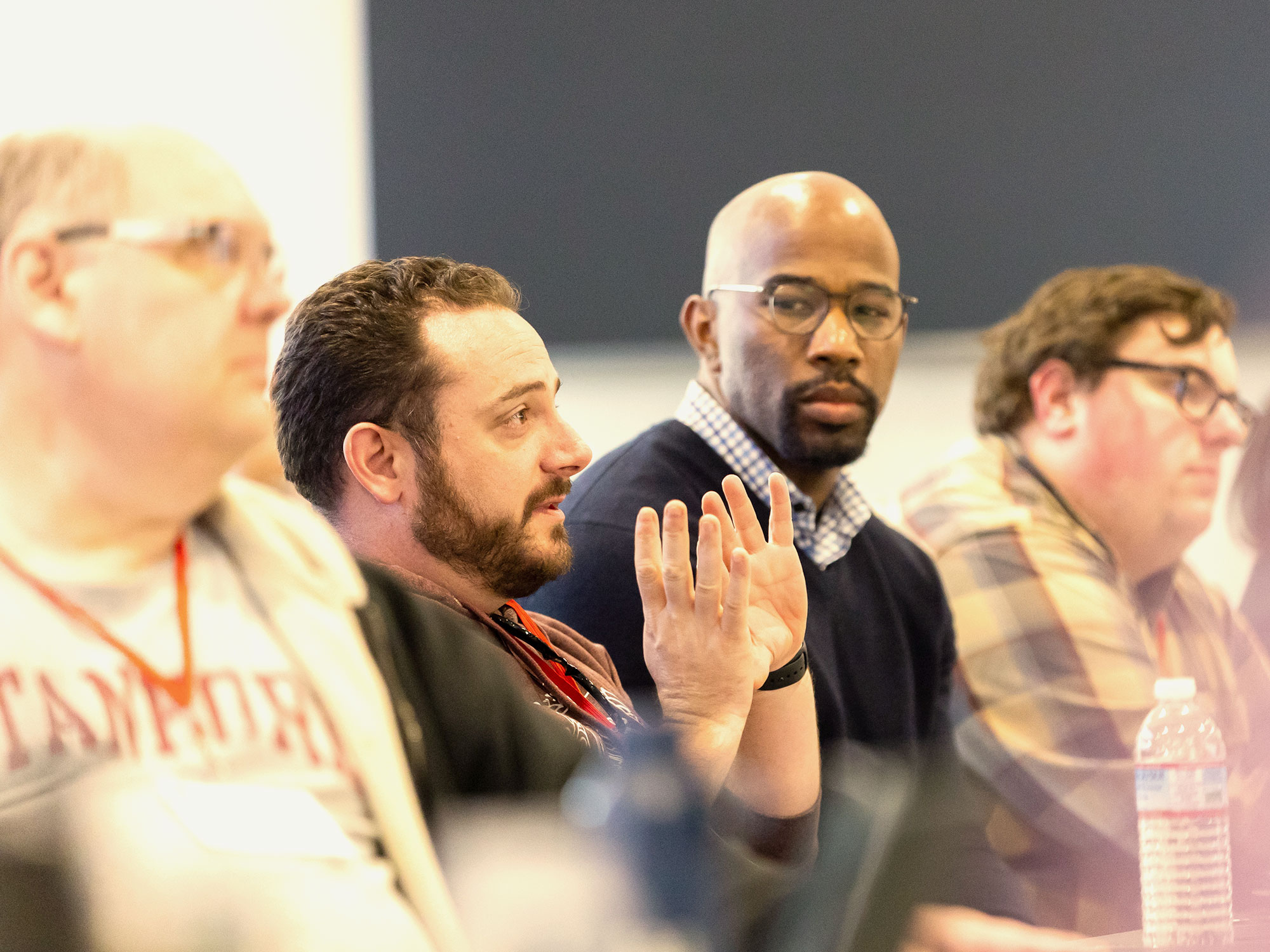 A man in a gray sweater addresses the group while others listen, including a man in glasses beside him.