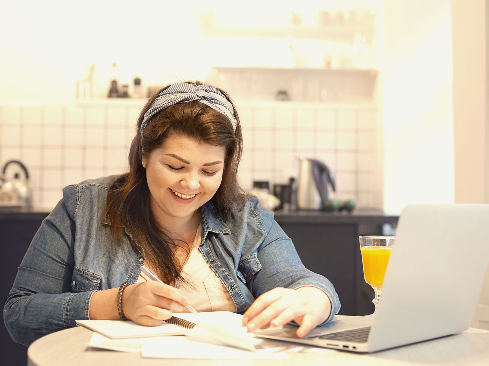 A smiling woman takes notes in front of a laptop at a kitchen table.