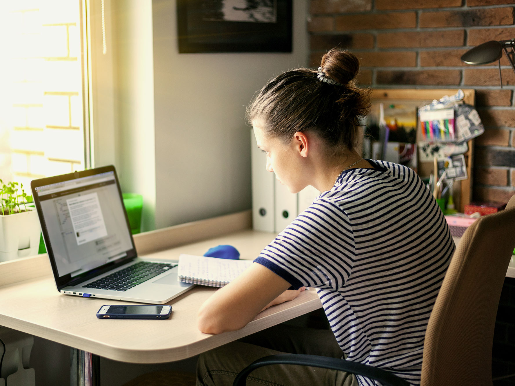 A woman sits at a desk while taking notes in front of a laptop.