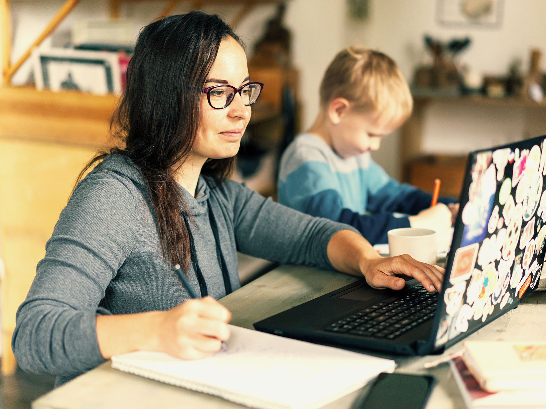 A woman types at a desk while taking notes. Her son sits next to her, writing on some paper.